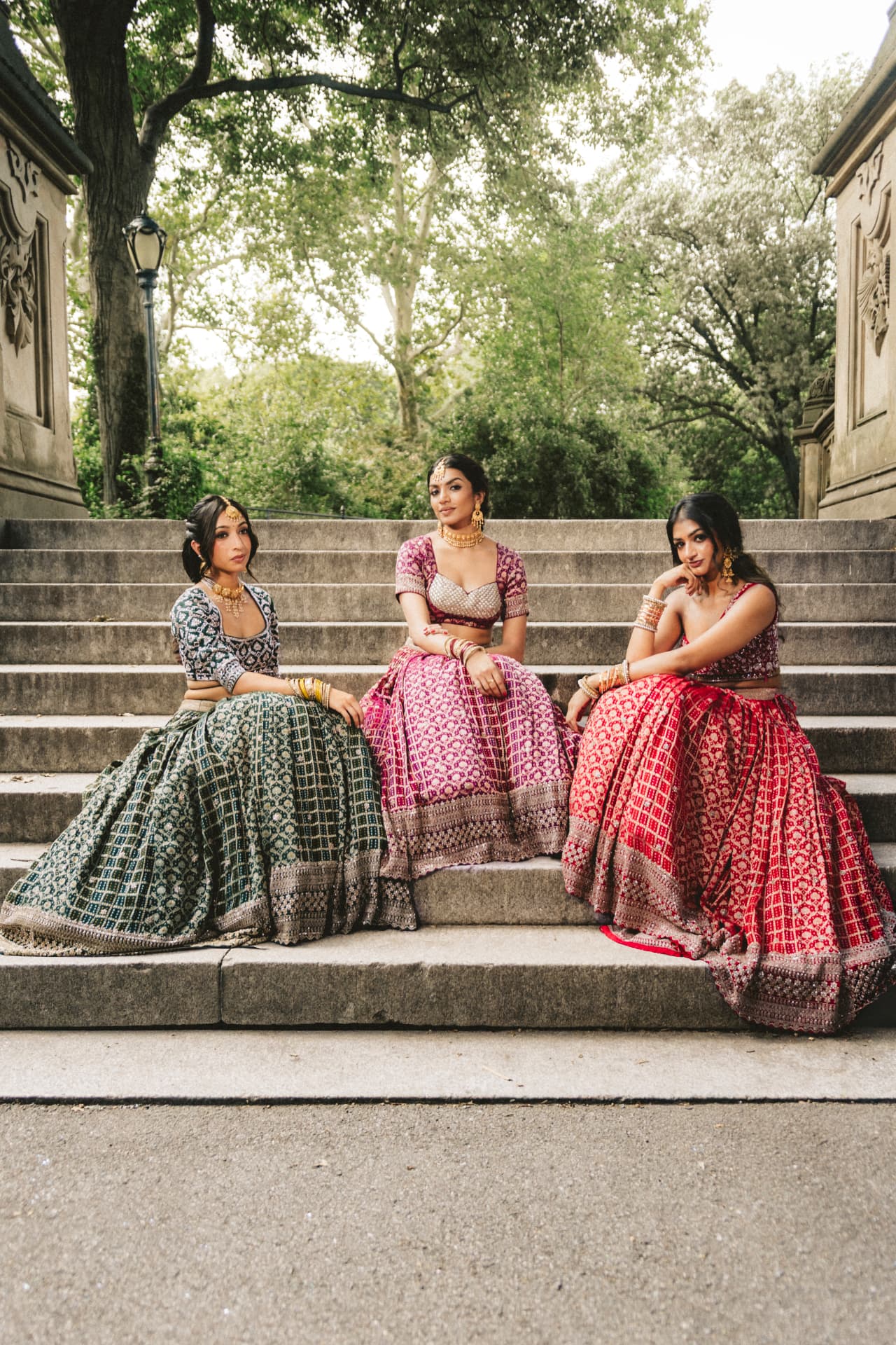 Three models seated on stone steps in emerald, magenta and red lehengas