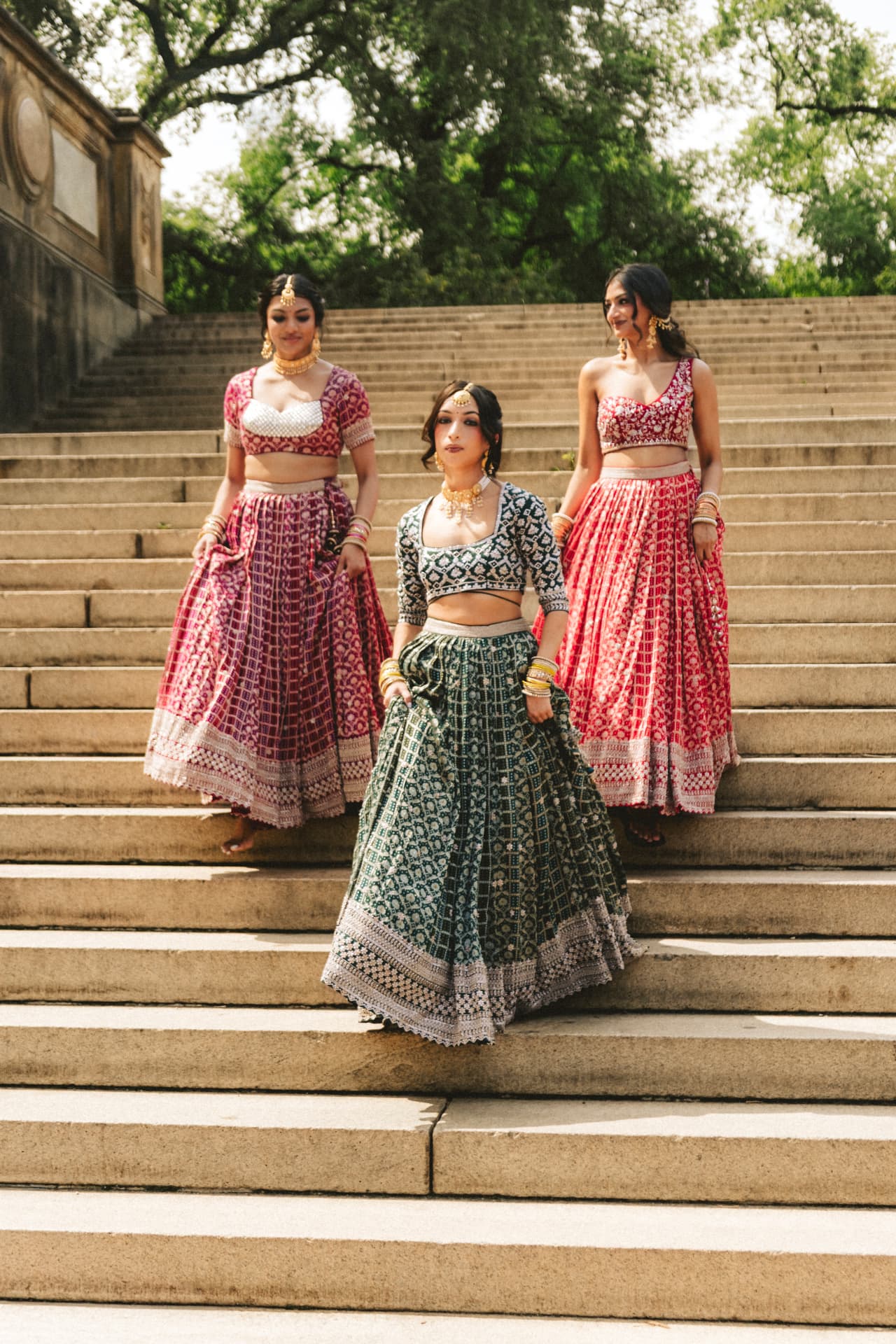 Three models descending stone steps in emerald, magenta and red lehengas